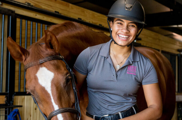 Female equestrian grins next to her brown horse in a stable