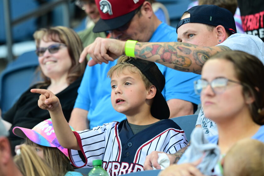 Young boy and his father point with excitement from the bleachers of a baseball game.