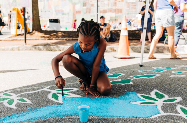 Black child painting vines and blue with chalk paint on a street