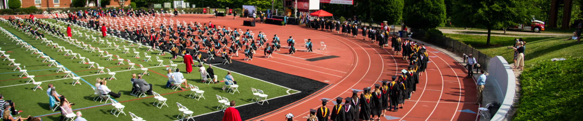 College graduates walk to their seats on a track and football field on graduation day.