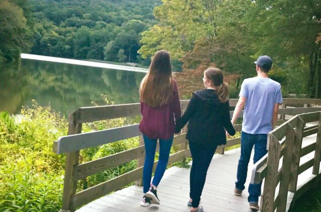 Family of three walks across a bridge, water, trees and a peak in the background
