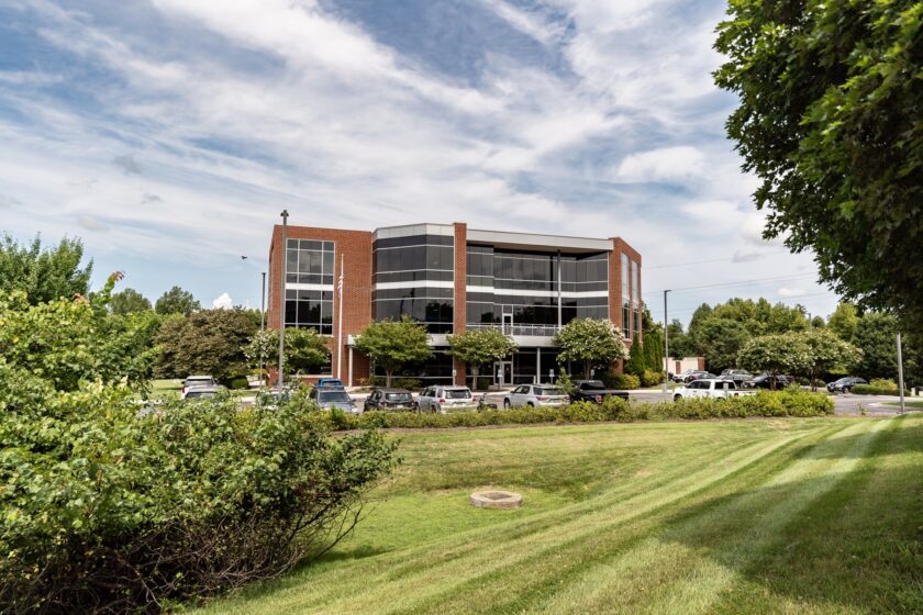 A modern three-story brick and glass office building surrounded by trees and a landscaped lawn under a partly cloudy sky