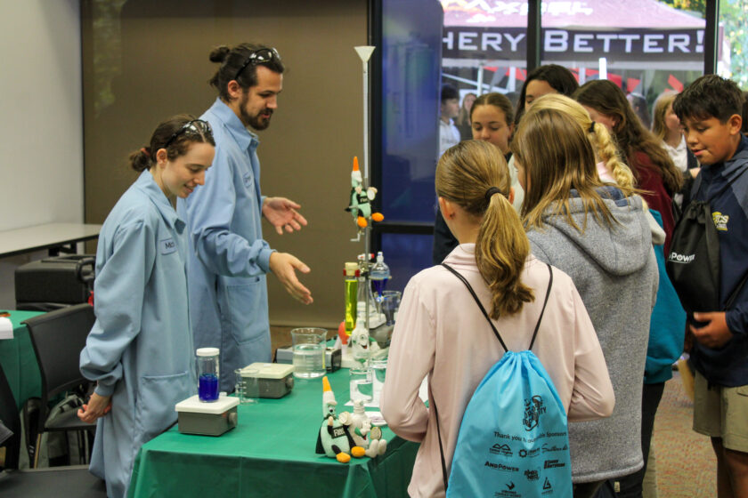 Students watch as two professionals in lab coats conduct an experiment at an exhibit table