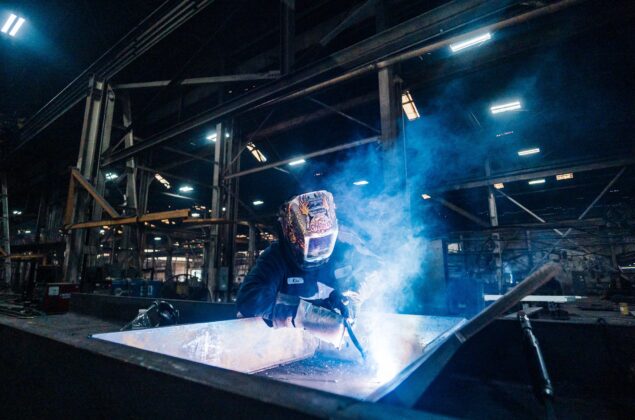 A welder with a mask works on metal in a lab.