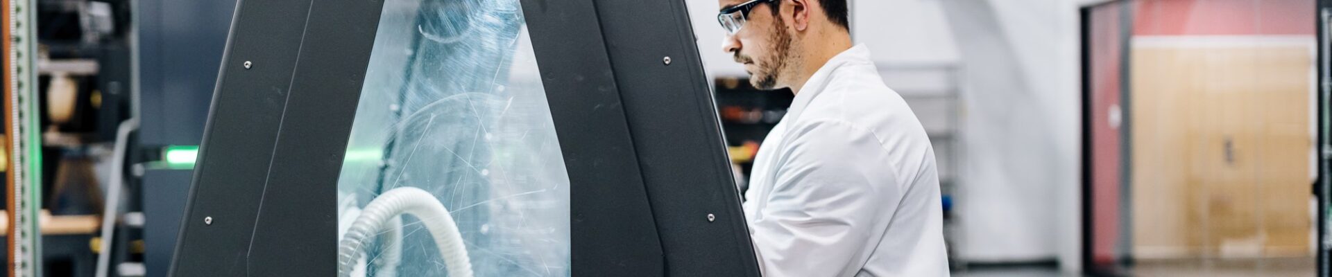 A technician in a white lab coat and safety glasses operates an enclosed industrial machine in a laboratory or manufacturing facility