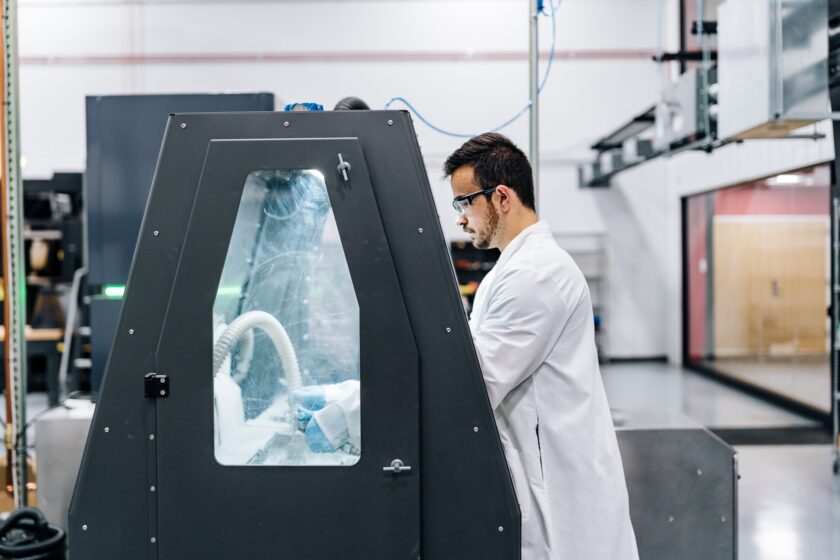 A technician in a white lab coat and safety glasses operates an enclosed industrial machine in a laboratory or manufacturing facility
