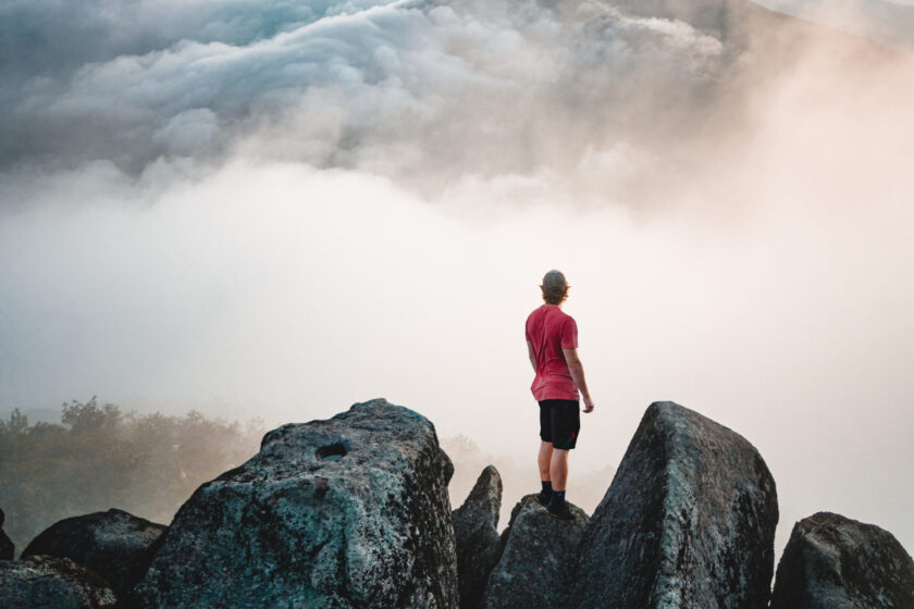 Person stands atop craggy mountain amidst clouds