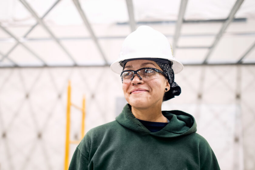 Female construction worker in a hard hat and safety glasses smiling at a job site.