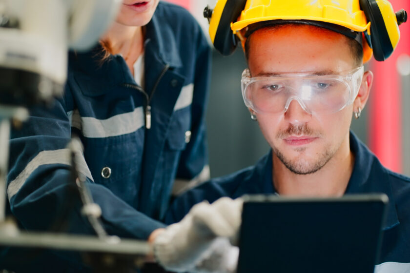 Technician in safety goggles and a hard hat using a tablet next to machinery.
