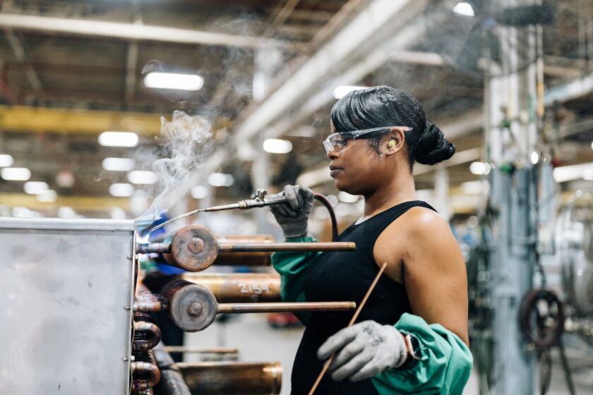 A worker in safety glasses and gloves uses a torch to braze copper components on a factory production floor