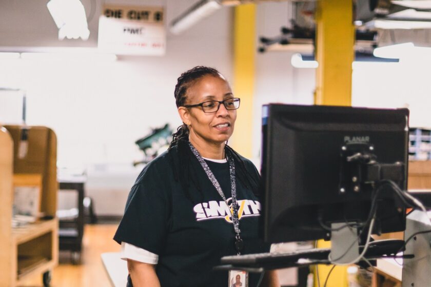 A smiling worker in glasses stands at a computer monitor on a production floor