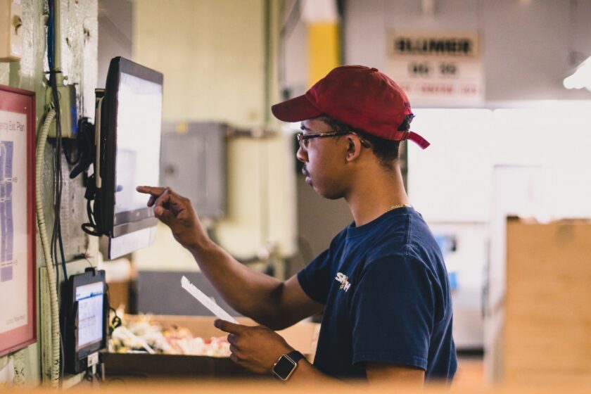 A young worker in a red cap points at a wall-mounted screen while holding a printout in a warehouse or production facility