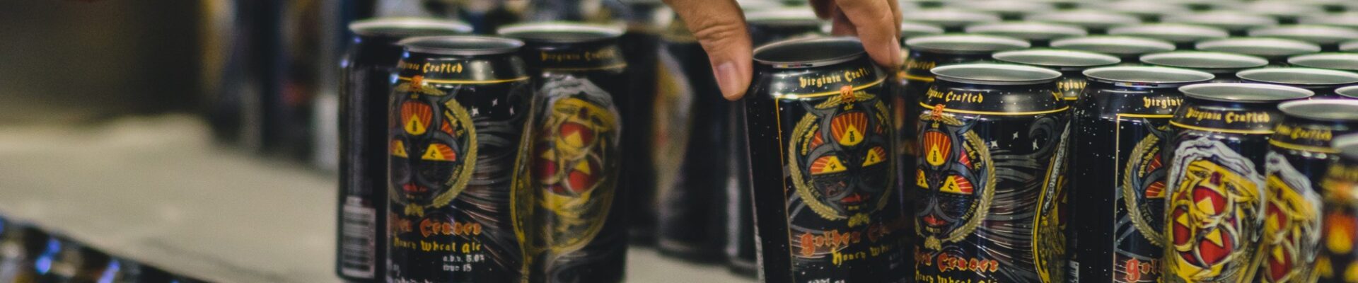 A hand reaches for a can on a packaging line filled with rows of dark, intricately designed craft beer cans