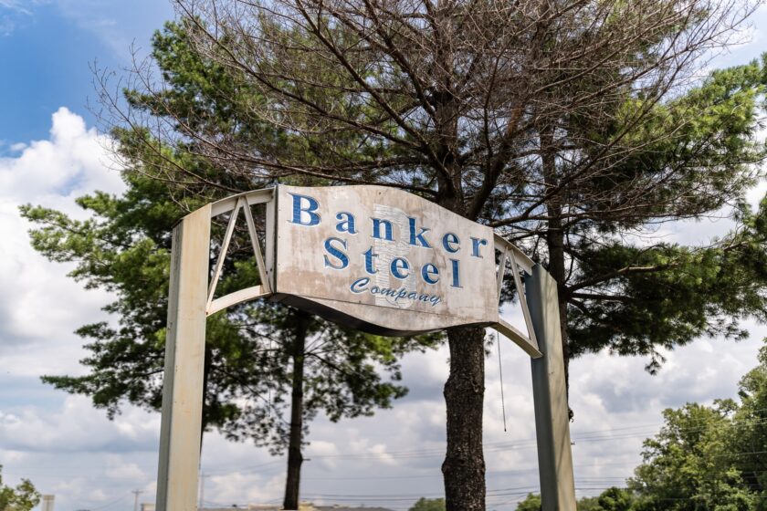 A weathered Banker Steel Company sign mounted on a metal frame against a backdrop of trees and a cloudy sky