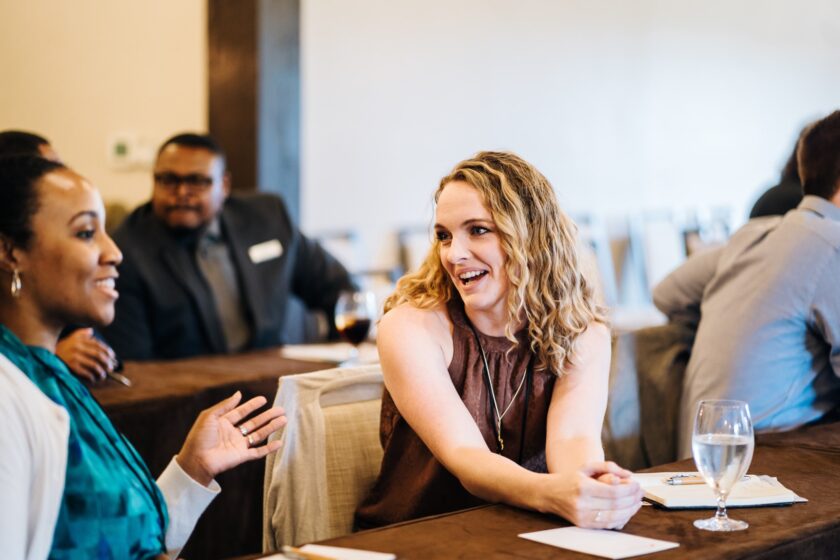 Two women smile and converse at a table during a networking or social event, with other attendees in the background