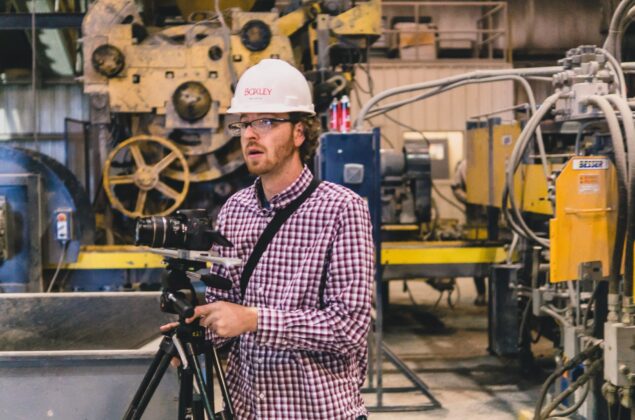 Man in a hard hat and safety glasses operating a tripod-mounted camera inside an industrial facility with heavy machinery.