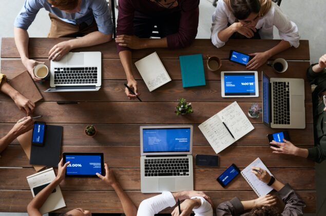 A group of coworkers sit together at a table strewn with notebooks and laptops.