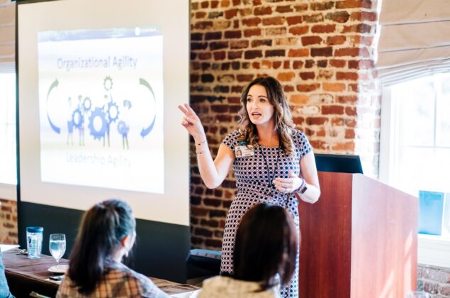 A woman gives a presentation on organizational and leadership agility to a seated audience, with a projector screen and exposed brick wall behind her