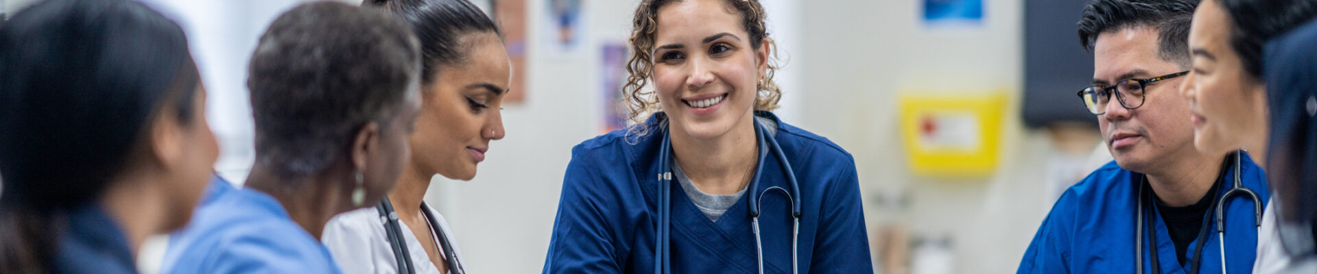 A small group of medical professionals gather around a table as they meet together to discuss patient cases. They are each dressed professionally and have files scattered between them for reference.