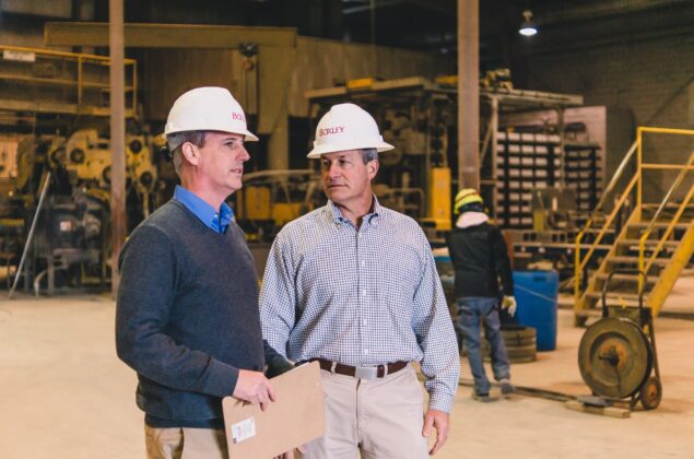 Two men in white hard hats stand and talk inside an industrial facility, with machinery and a worker in the background
